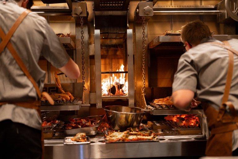 Two chefs working at an open wood-fired grill, cooking seafood and other dishes over glowing embers.