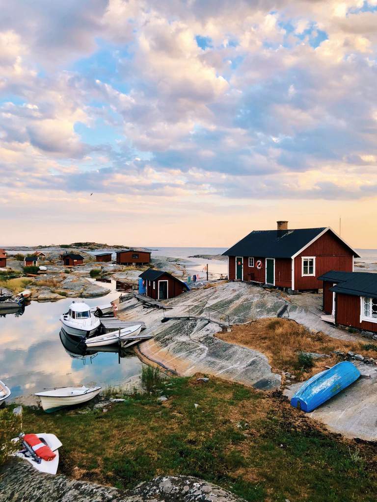 Small red cottages and boats on the rocky shore of a quiet bay in the Stockholm archipelago under a cloudy evening sky with soft light.