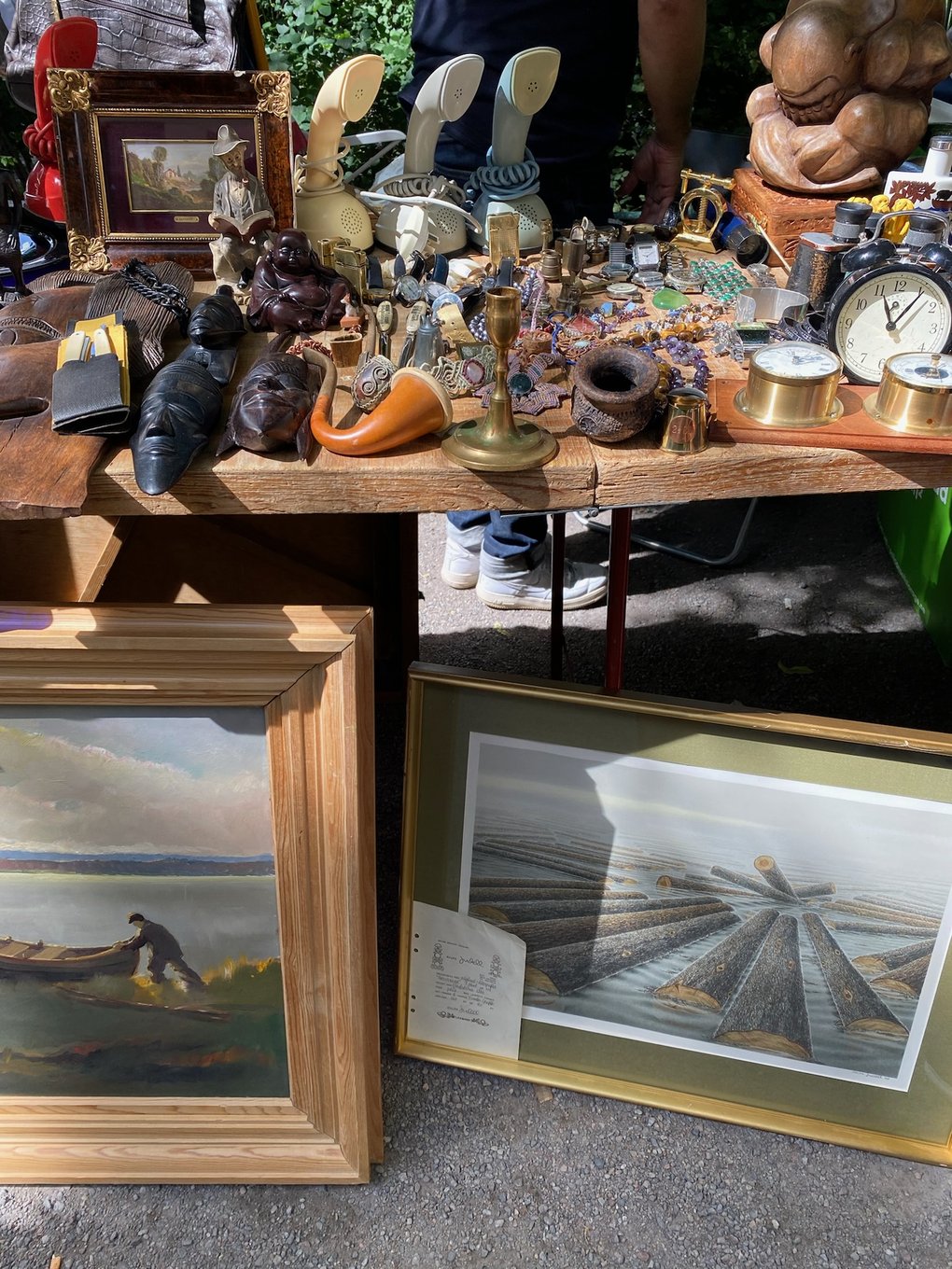 A flea market table filled with assorted vintage items, including small statues, clocks, pipes, and brass objects, with framed paintings and prints leaning against the table.