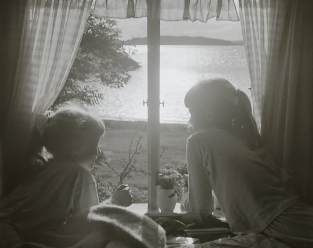 Two girls with their backs to the viewer look out a window towards a shimmering lake in the sunlight. A poetic photograph from the Nordic Museum’s collections.