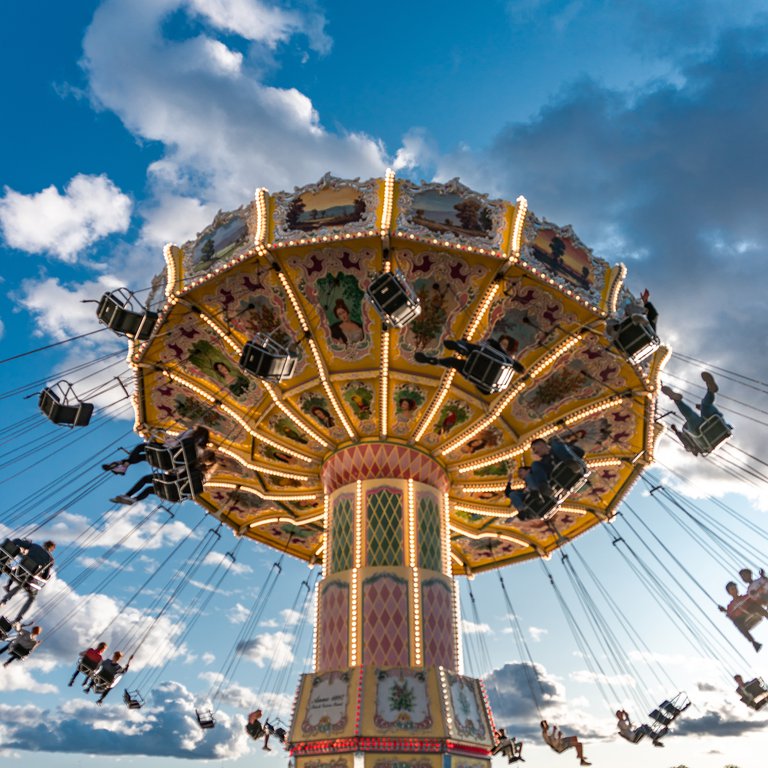People enjoy a colorful swing carousel ride at an amusement park under a bright blue sky with scattered clouds.